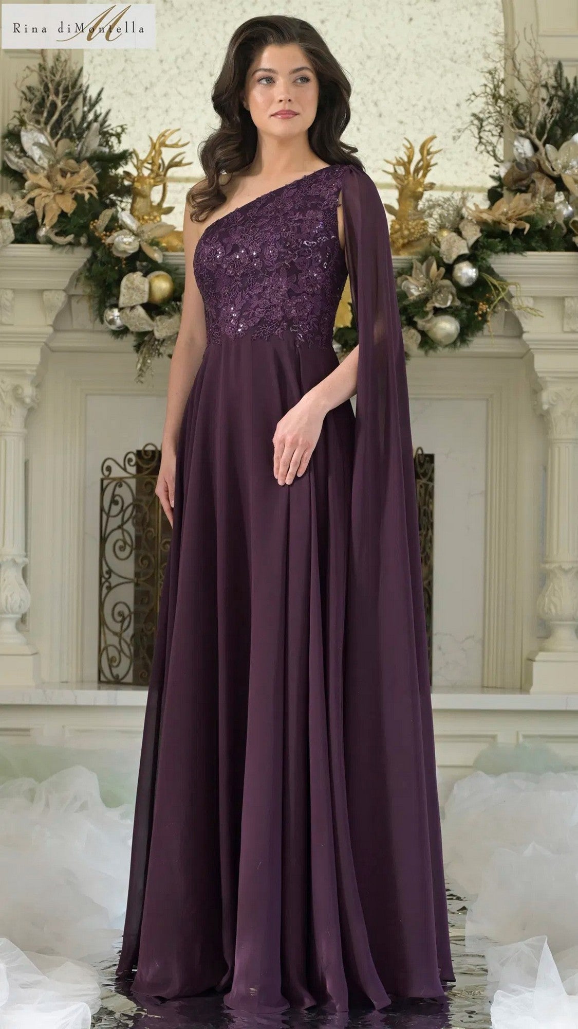 A woman stands indoors before a decorated mantel, wearing the Long Formal Dress: Rina Di Montella RD3354 in eggplant—a floor-length, one-shoulder gown featuring a floral-embroidered bodice.